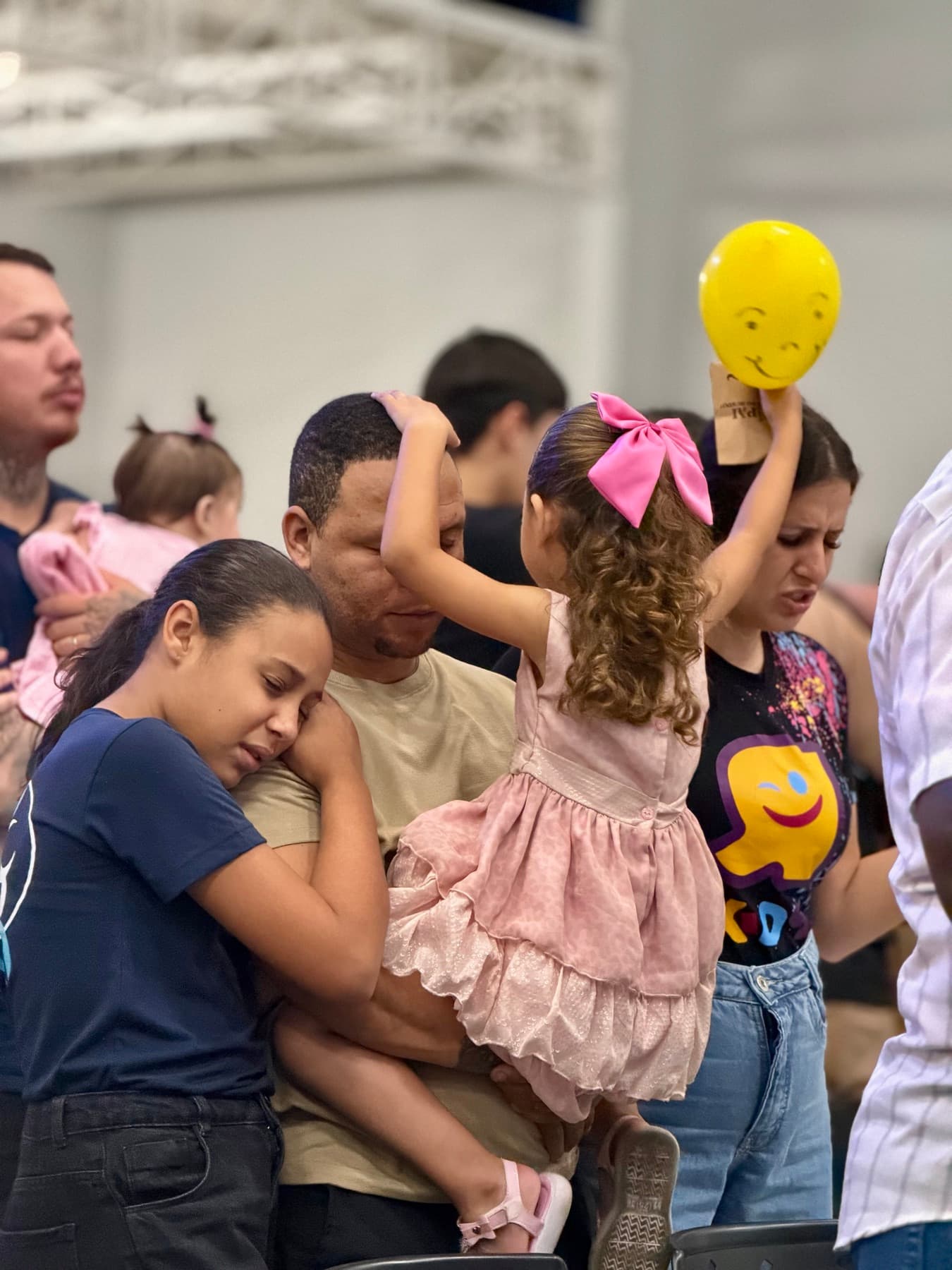 Pai com as duas filhas no colo durante um culto, uma delas com a cabeça apoiada no ombro dele