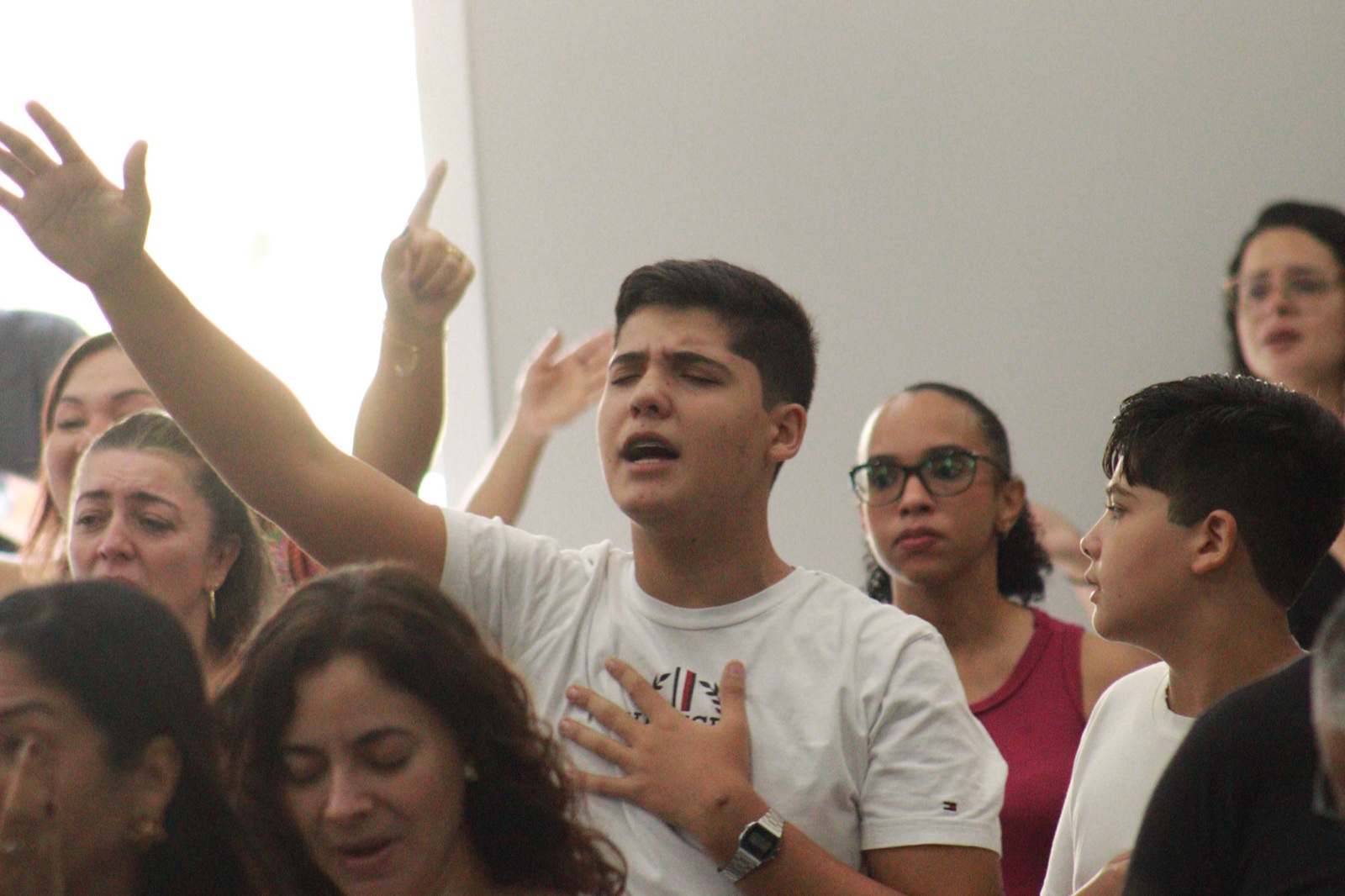 Jovem de camiseta branca cantando de olhos fechados no meio da congregação, com um braço erguido e a outra mão sobre o peito