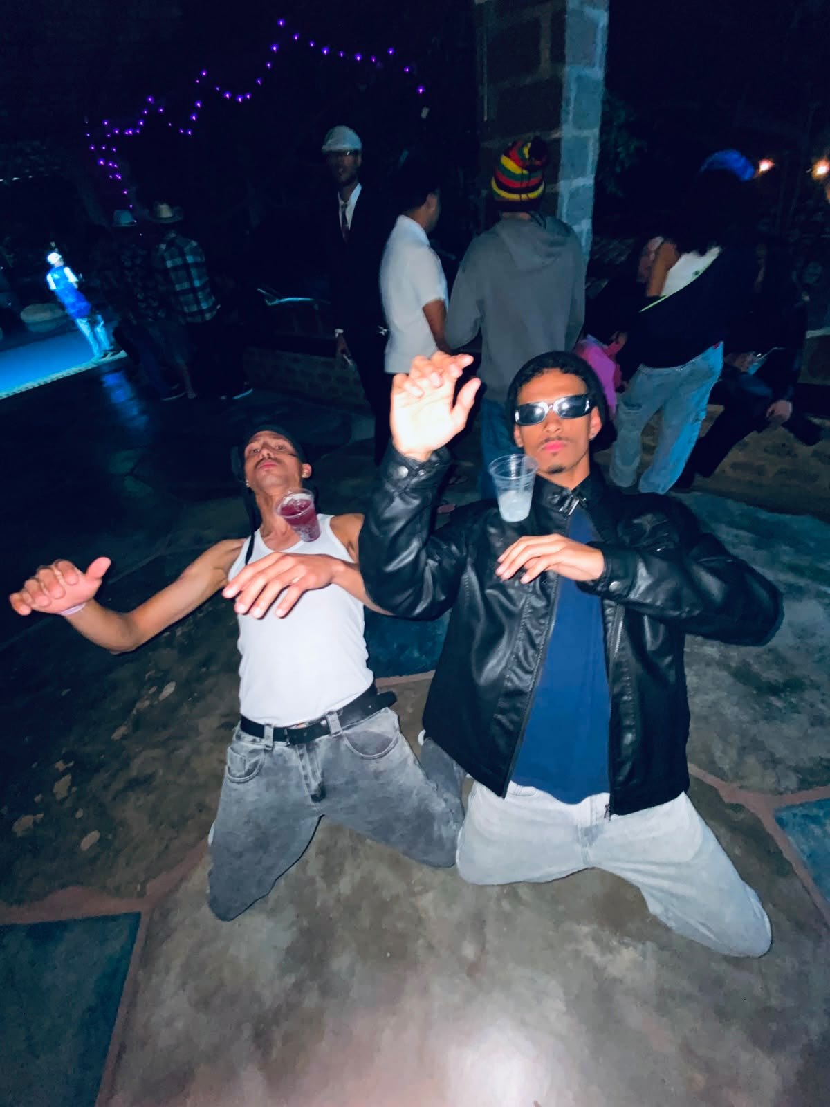 Dois rapazes ajoelhados de braços abertos numa festa à noite, com fumaça e luzes atrás