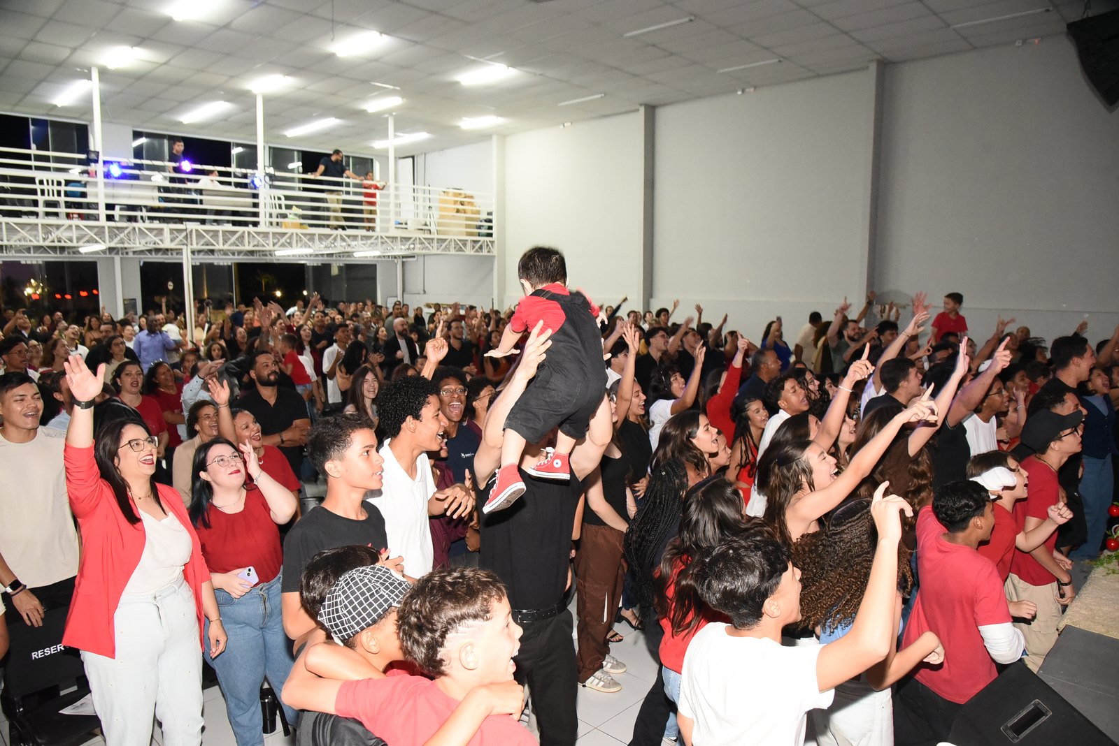 Salão lotado visto de trás, com dezenas de jovens de pé e de mãos erguidas