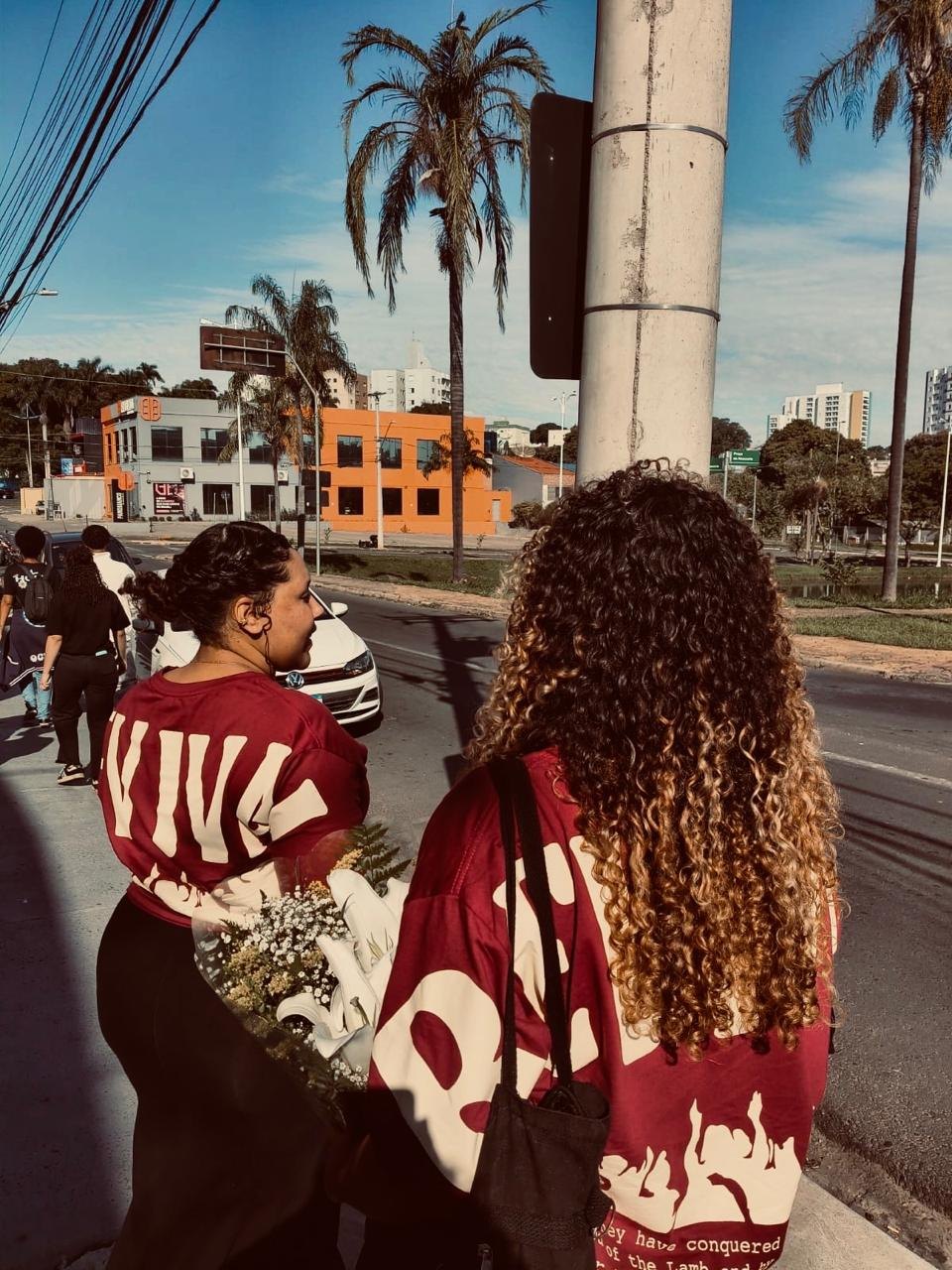 Duas jovens de costas, de jaqueta, paradas na esquina de uma avenida com palmeiras