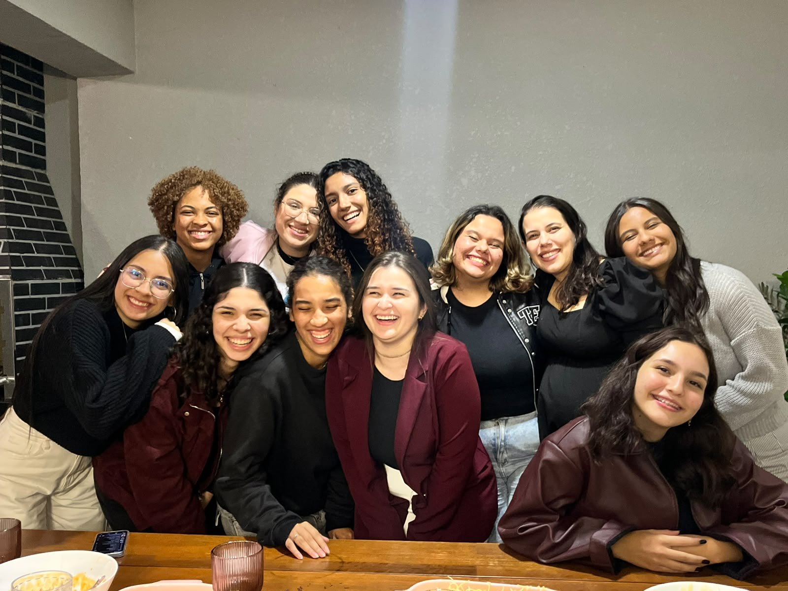 Roda de moças posando abraçadas na sala, algumas sentadas à mesa de madeira