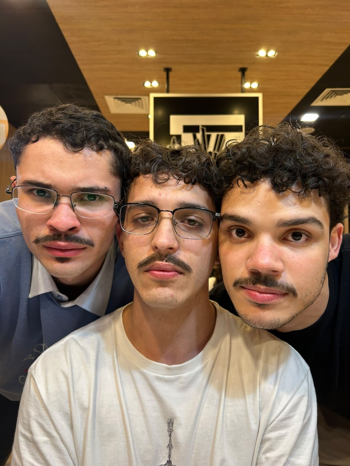 Três rapazes de bigode postiço posando sérios para a selfie dentro de um shopping