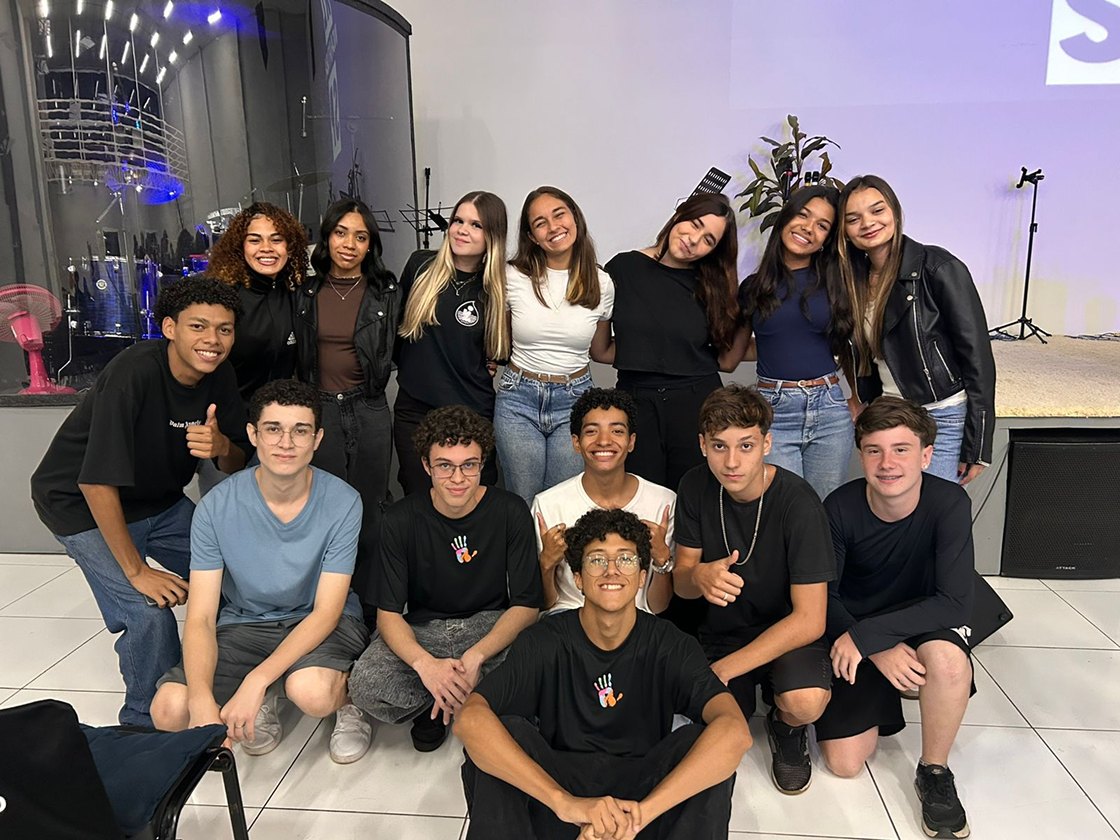Turma posando num salão com a parede lavada de luz roxa e o acrílico da bateria atrás