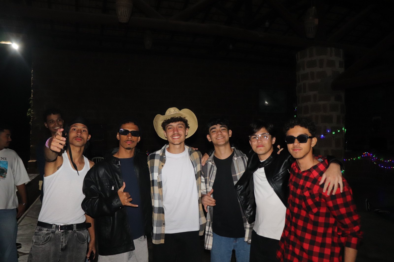 Um grupo de rapazes posando abraçados, ombro a ombro, numa varanda à noite — um deles de chapéu de palha, dois de óculos escuros —, com luzinhas coloridas acesas na parede ao fundo