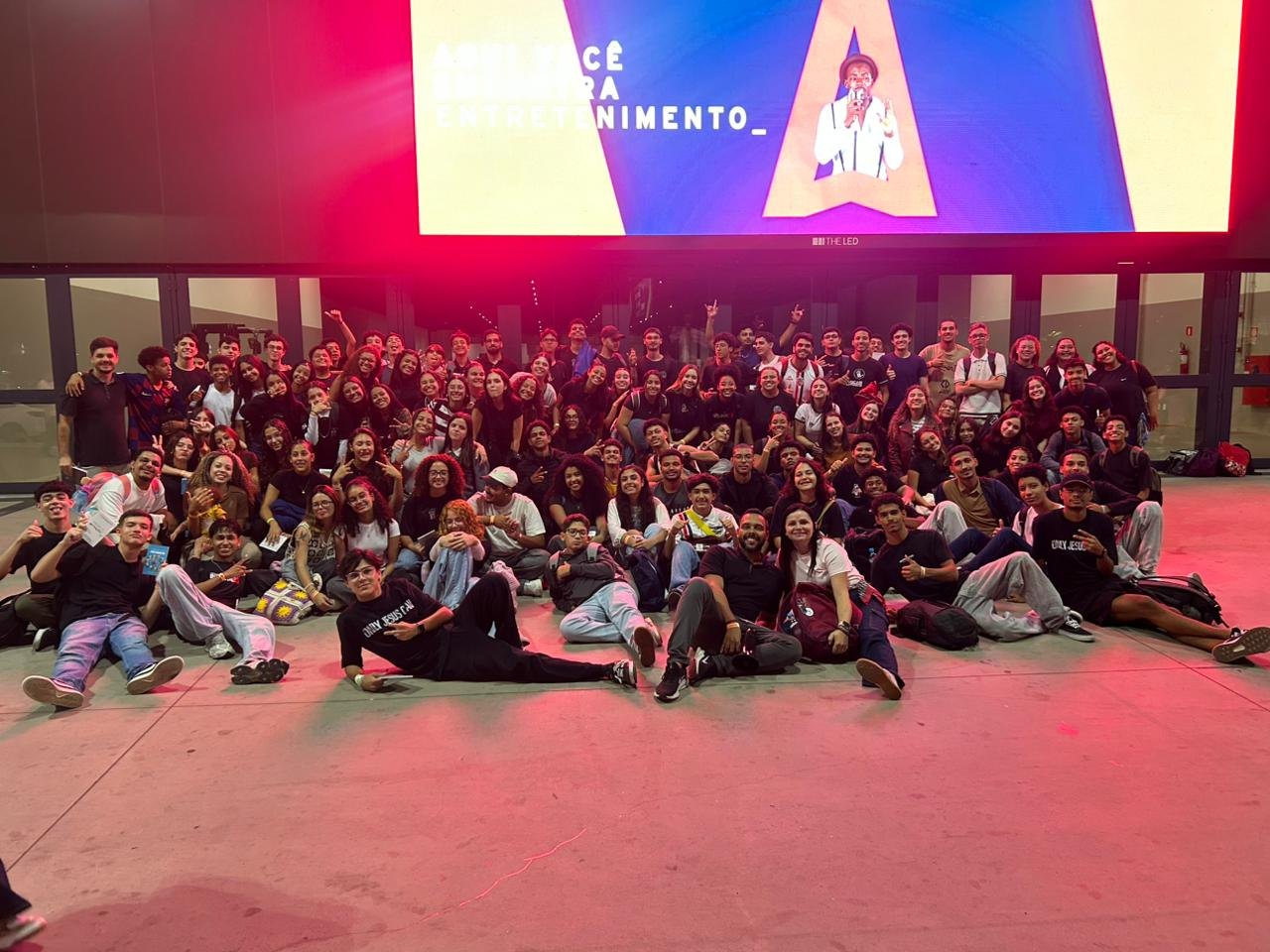 Foto de turma com mais de cem jovens sentados e em pé diante do telão, o salão inteiro tomado por uma luz rosa